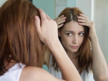 Porträt einer schönen jungen Frau, die ihre Kopfhaut und Haare vor dem Spiegel, Haarwurzeln, Farbe, grauehaare, Haarausfall oder trockene Kopfhaut Problem untersucht | © Getty Images/triocean