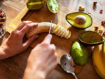 Frau schneidet auf einem Holzbrett Banane und Avocado. | © Getty Images / Marko Geber