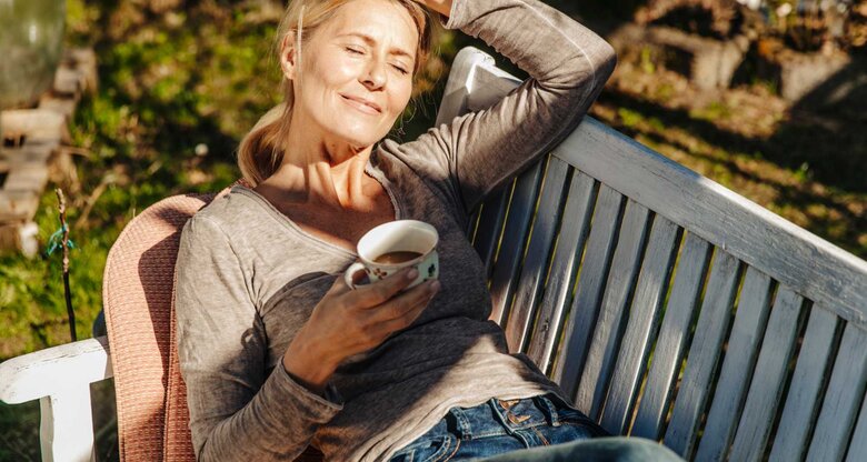 Frau liegt mit einer Tasse Kaffee im Garten auf einer Bank und entspannt sich. | © Getty Images/Westend61