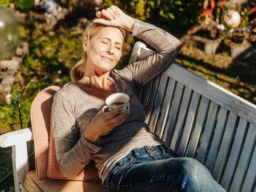 Frau liegt mit einer Tasse Kaffee im Garten auf einer Bank und entspannt sich. | © Getty Images/Westend61