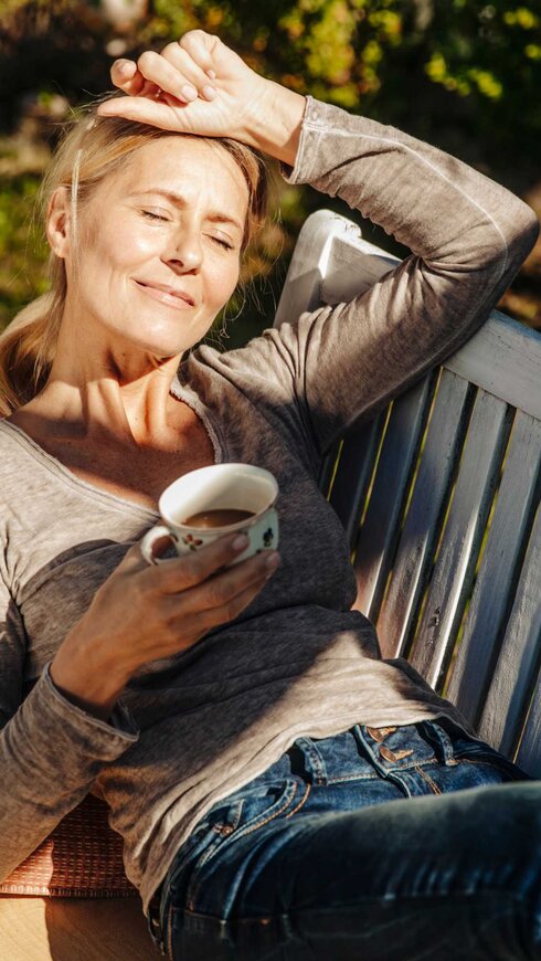 Frau liegt mit einer Tasse Kaffee im Garten auf einer Bank und entspannt sich.