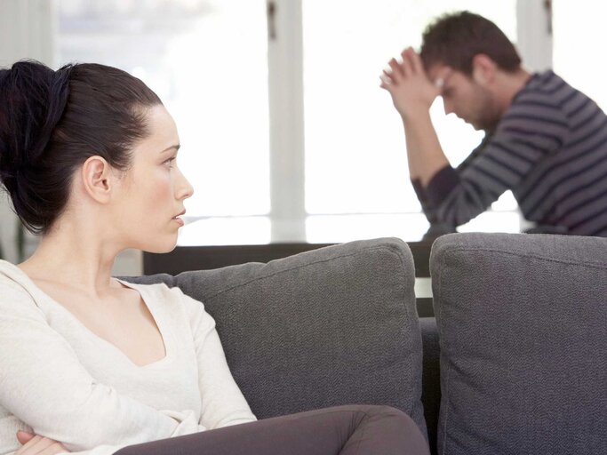 Frau sitzt auf der Couch und blickt zu ihrem Partner, der nachdenklich am Tisch sitzt | © Getty Images/Noel Hendrickson