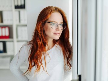 Junge Frau mit roten Haaren und Brille schaut nachdenklich aus dem Fenster | © Getty Images/stockfour