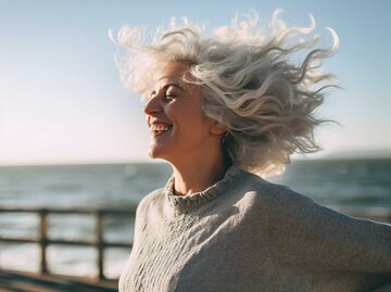 Glückliche Frau steht am Strand mit Wind im Haar | © Midjourney/Maxi Baumgärtner