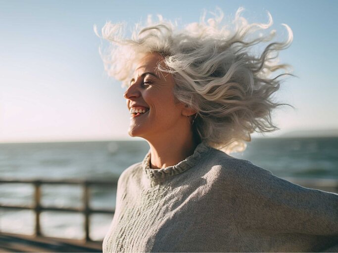 Glückliche Frau steht am Strand mit Wind im Haar | © Midjourney/Maxi Baumgärtner