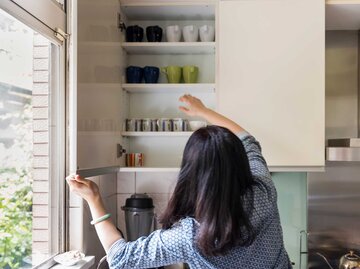 Frau mit dunklen Haaren öffnet einen Küchenschrank, in dem Tassen stehen. | © Getty Images / Leren Lu