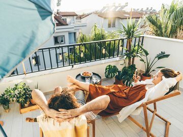 Eine Frau liegt auf einem Liegestuhl auf ihrem Balkon inmitten von Pflanzen | © GettyImages/Maria Korneeva