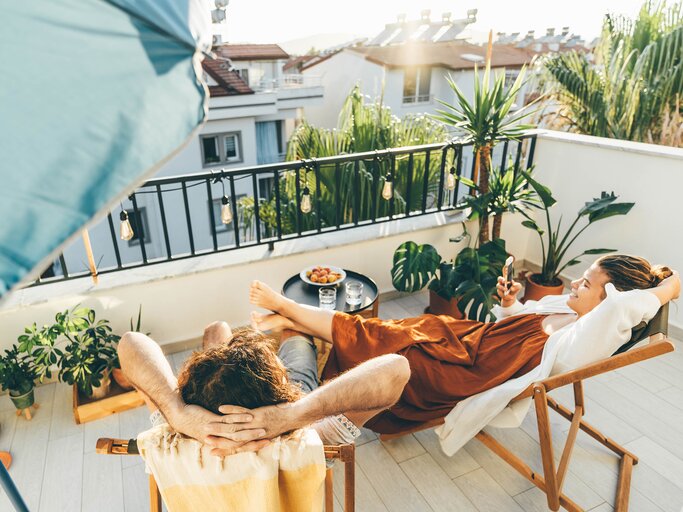 Eine Frau liegt auf einem Liegestuhl auf ihrem Balkon inmitten von Pflanzen | © GettyImages/Maria Korneeva