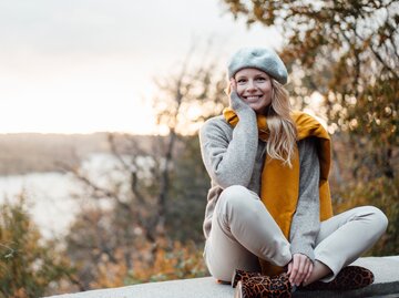 Frau sitzt im Schneidersitz auf Stützmauer und trägt modisches Herbstoutfit | © GettyImages/Westend61