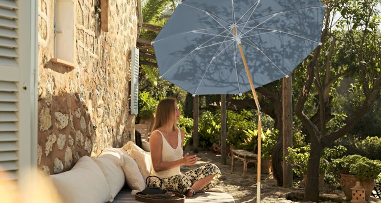 Frau sitzt in mediterranem Garten unter einem Sonnenschirm | © Glatz