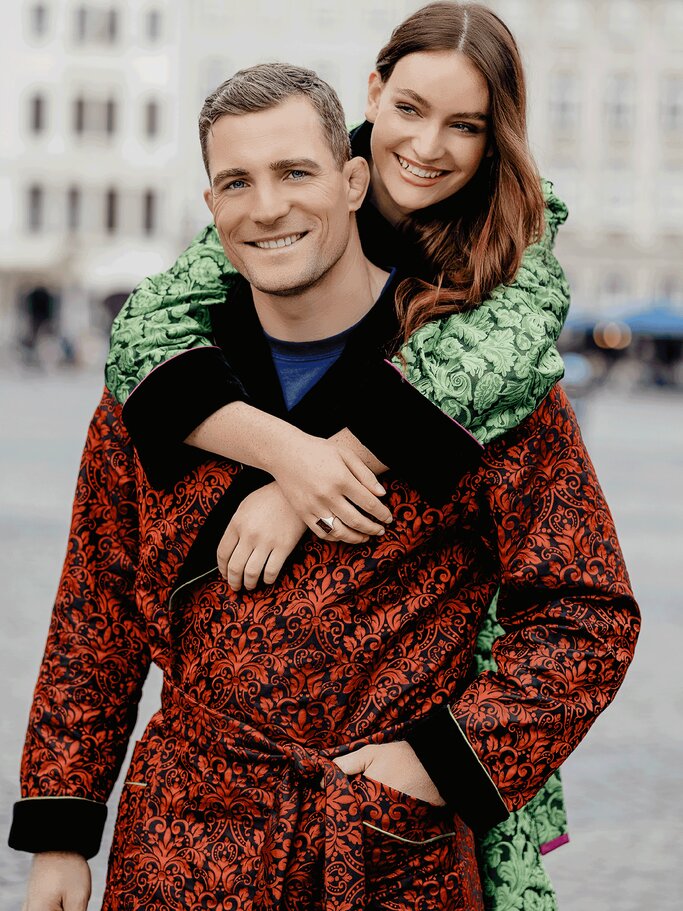 Junger Mann in rot-schwarzem Jacquard-Mantel lächelt, während ihn eine junge Frau im grünen Mantel von hinten umarmt. | © Wagler Couture
