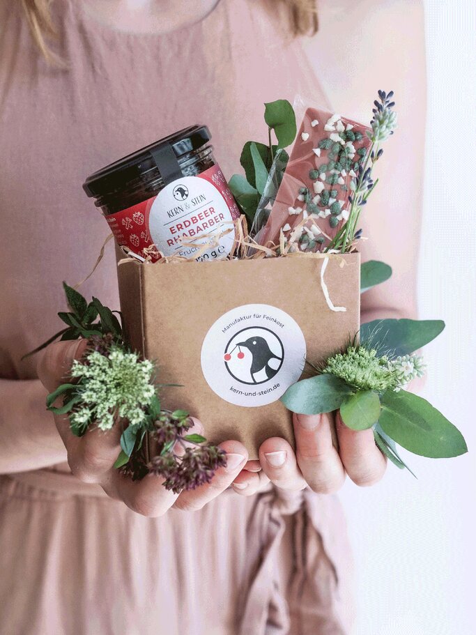 Junge Frau in rosa Kleid hält eine Geschenkbox mit Erdbeer-Rhabarber-Konfitüre, rosa Schokolade und frischen Blumen. | © Kern und Stein