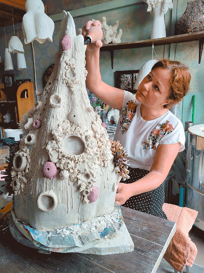 Eine rothaarige Frau mittleren Alters verziert konzentriert eine hohe, organisch geformte Porzellanskulptur in einem Atelier. | © Stefan Passig