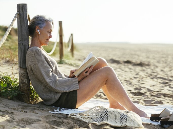Reife Frau liegt am Strand und liest ein Buch | © GettyImages/Westend61