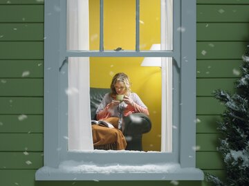 Frau liest vor dem Fenster ein Buch, draußen liegt Schnee, gemütliche Atmosphäre.  | © GettyImages/	Stephen Swintek