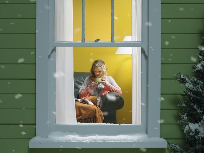 Frau liest vor dem Fenster ein Buch, draußen liegt Schnee, gemütliche Atmosphäre.  | © GettyImages/	Stephen Swintek