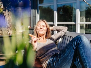 Frau liegt auf einer Bank im Garten in der Sonne uns isst eine Karotte. | © Getty Images/Westend61