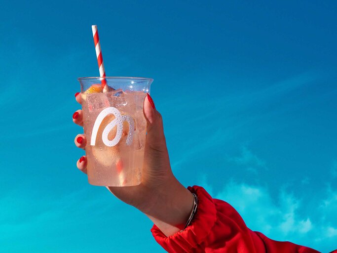 Hand hält einen Becher mit FIZI Limonade in die Luft, im Hintergrund ein blauer Himmel. | © More Nutrition