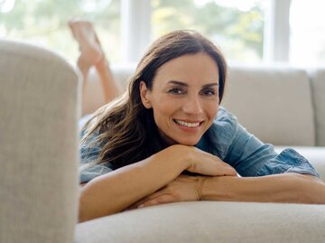 Schöne Frau im höheren Alter liegt bäuchlings auf der Couch und lächelt in die Kamera. | © Getty Images / Westend61