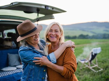 Zwei Frauen stehen vor ihrem Campingwagen im Grünen und umarmen sich fröhlich. | © Getty Images / Halfpoint Images