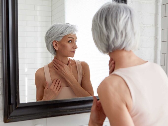 Frau mit grauen Haaren betrachtet sich im Spiegel und begutachtet ihren Hals. | © Getty Images / Ekaterina Demidova