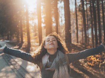 Eine Frau breitet ihre Arme aus und steht an einem goldenen Oktobertag im Wald | © GettyImages/Milan Markovic