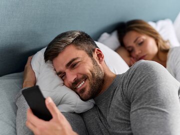 Mann benutzt Handy und checkt Nachrichten im Bett, während seine Frau schläft | © GettyImages/EmirMemedovski