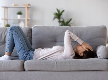 Eine Frau liegt auf der Couch und hält sich die Hände vors Gesicht | © GettyImages/fizkes