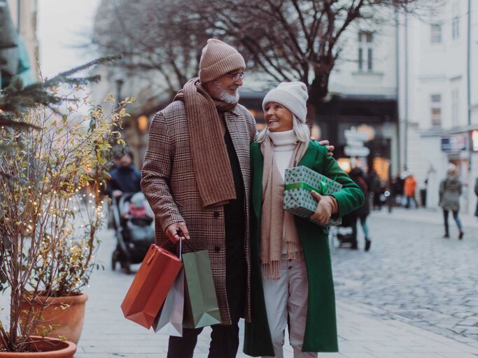 Älteres Paar schlendert Arm in Arm mit Geschenken zur Weihnachtszeit durch die Einkaufsstraße. | © Adobe Stock/Halfpoint