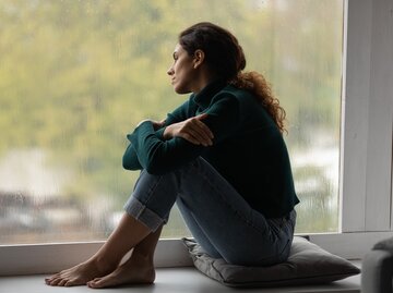 Eine Frau sitzt auf dem Fenstersims und schaut nachdenklich aus dem Fenster | © GettyImages/fizkes