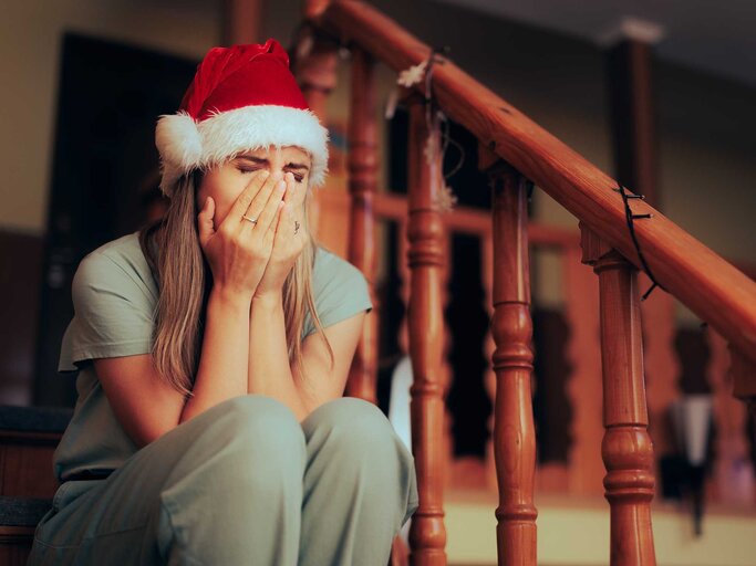 Frau sitzt auf der Treppe und trägt eine Weihnachtsmütze und hat ihr Gesicht in den Händen vergraben. | © Adobe Stock/nicoletaionescu
