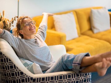 Frau sitzt auf einem Wohnzimmersessel und streckt entspannt und glücklich die Arme in die Luft. | © Adobe Stock/simona