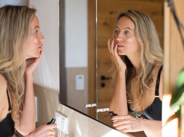 Blonde Frau steht vor dem Spiegel und trägt Foundation auf. | © Getty Images / Kathrin Ziegler
