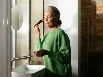 Frau mit grauen Haaren steht im Badezimmer und schminkt sich mit einem Pinsel. | © Getty Images / Westend61
