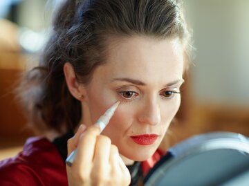 Frau sitzt vor Handspiegel und trägt Concealer auf | © Getty Images/CentralITAlliance