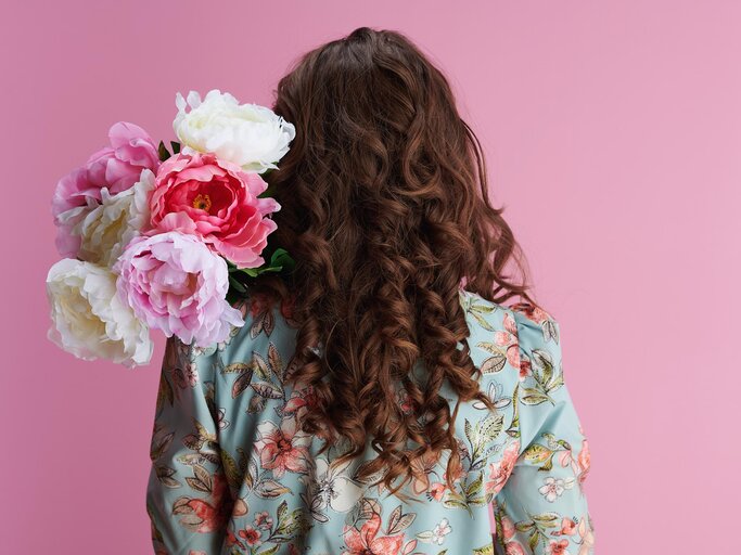 Rückenansicht von Frau mit braunen, voluminösen Haaren, die Pfingstrosen im Arm hält | © Getty Images/CentralITAlliance
