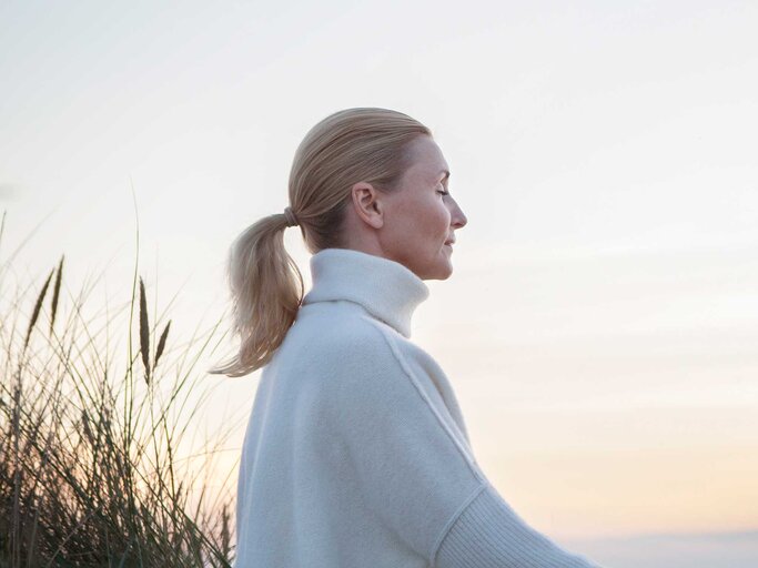 Frau trägt ihre Haare in einem Zopf und sitzt mit einem warmen Pullover am Strand. | © Getty Images / Henrik Sorensen