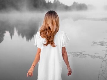 Frau mit langen, hellbraunen Haaren steht bei Nebel an See | © Getty Images/Azovsky