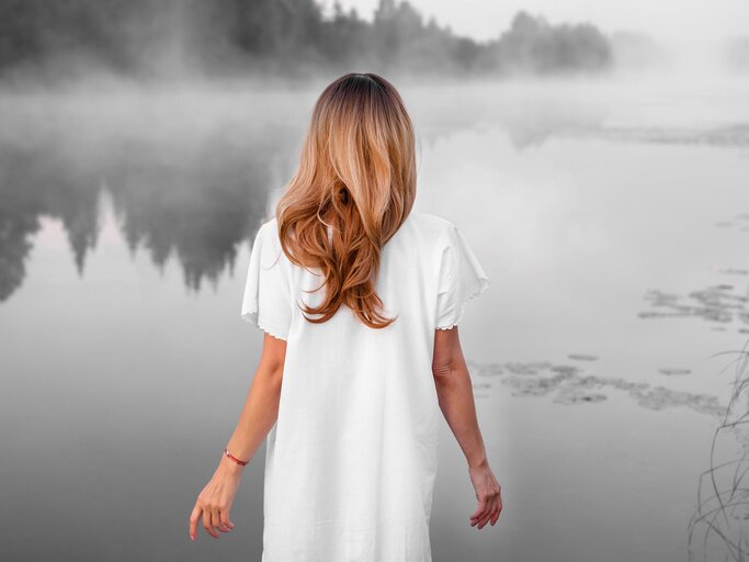 Frau mit langen, hellbraunen Haaren steht bei Nebel an See | © Getty Images/Azovsky