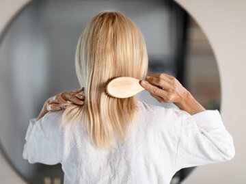 Frau mit blonden Haaren steht vor dem Spiegel und kämmt sich die Haare. | © Adobe Stock/Prostock-studio