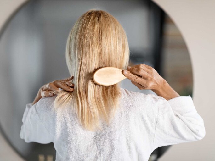 Frau mit blonden Haaren steht vor dem Spiegel und kämmt sich die Haare. | © Adobe Stock/Prostock-studio