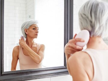 Frau mit kurzen grauen Haaren kämmt sich die Haare vor dem Spiegel | © Getty Images/Ekaterina Demidova