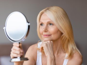 Frau mit blonden Haaren schaut in Handspiegel | © AdobeStock/Prostock-studio