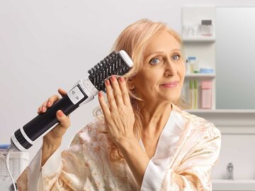 Frau steht im Badezimmer und stylt ihre Haare mit einer Warmluftbürste. | © Adobe Stock/Ljupco Smokovski