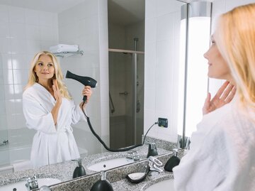 Frau steht mit Bademantel im Badezimmer und föhnt sich die Haare. | © Adobe Stock/LIGHTFIELD STUDIOS