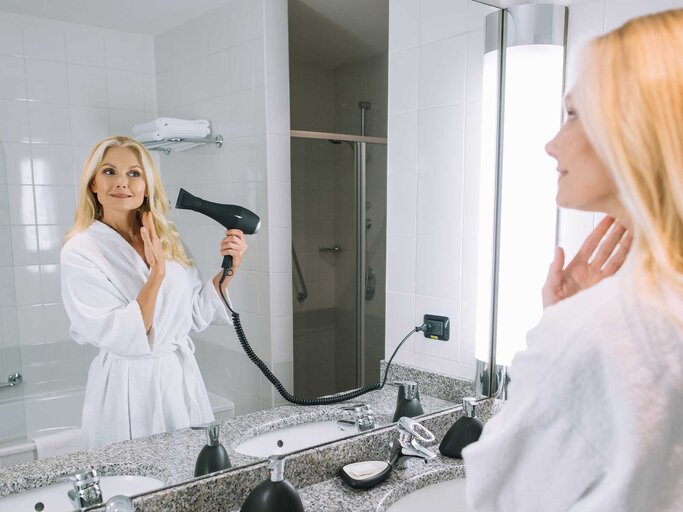 Frau steht mit Bademantel im Badezimmer und föhnt sich die Haare. | © Adobe Stock/LIGHTFIELD STUDIOS