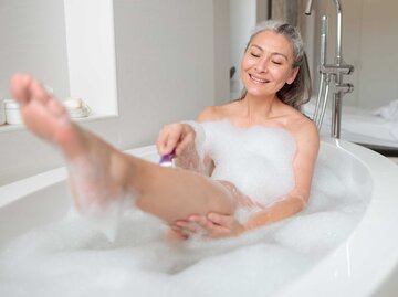 Asiatische Frau sitzt in der Badewanne und rasiert sich die Beine. | © Getty Images / Creative Credit