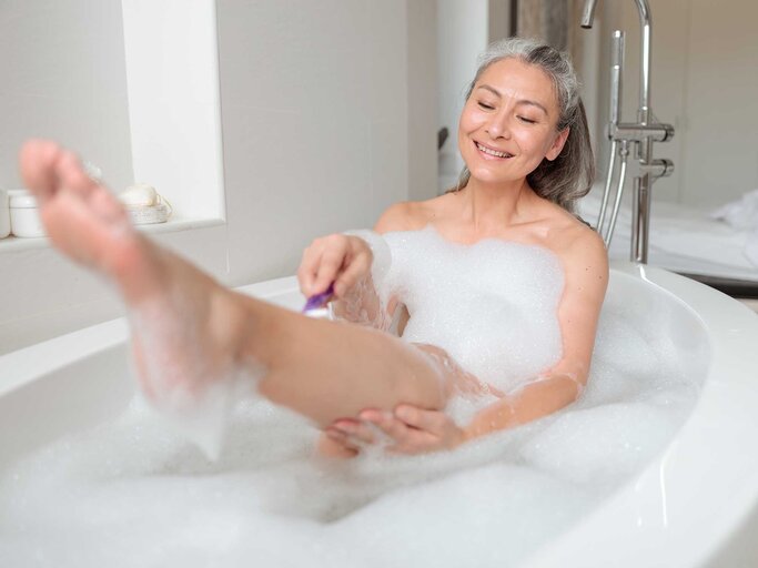 Asiatische Frau sitzt in der Badewanne und rasiert sich die Beine. | © Getty Images / Creative Credit