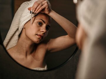Frau mit Handtuch in den Haaren steht vor dem Spiegel und trägt Serum im Gesicht auf. | © Adobe Stock / La Famiglia
