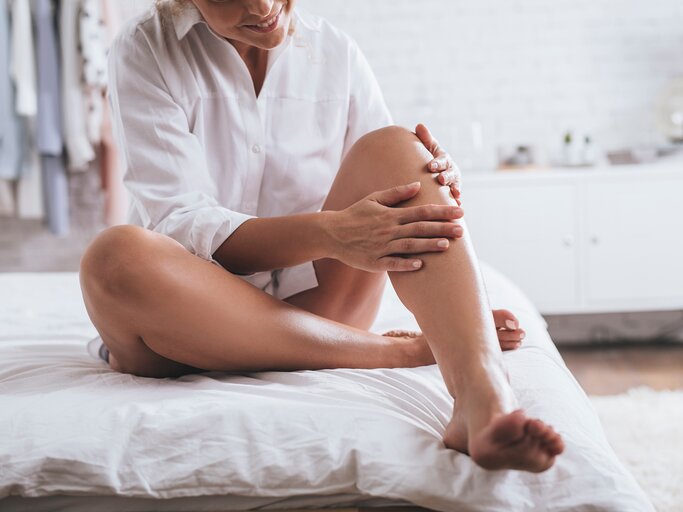 Eine Frau sitzt auf dem Bett und cremt sich die Beine ein | © Adobe Stock/LStockStudio